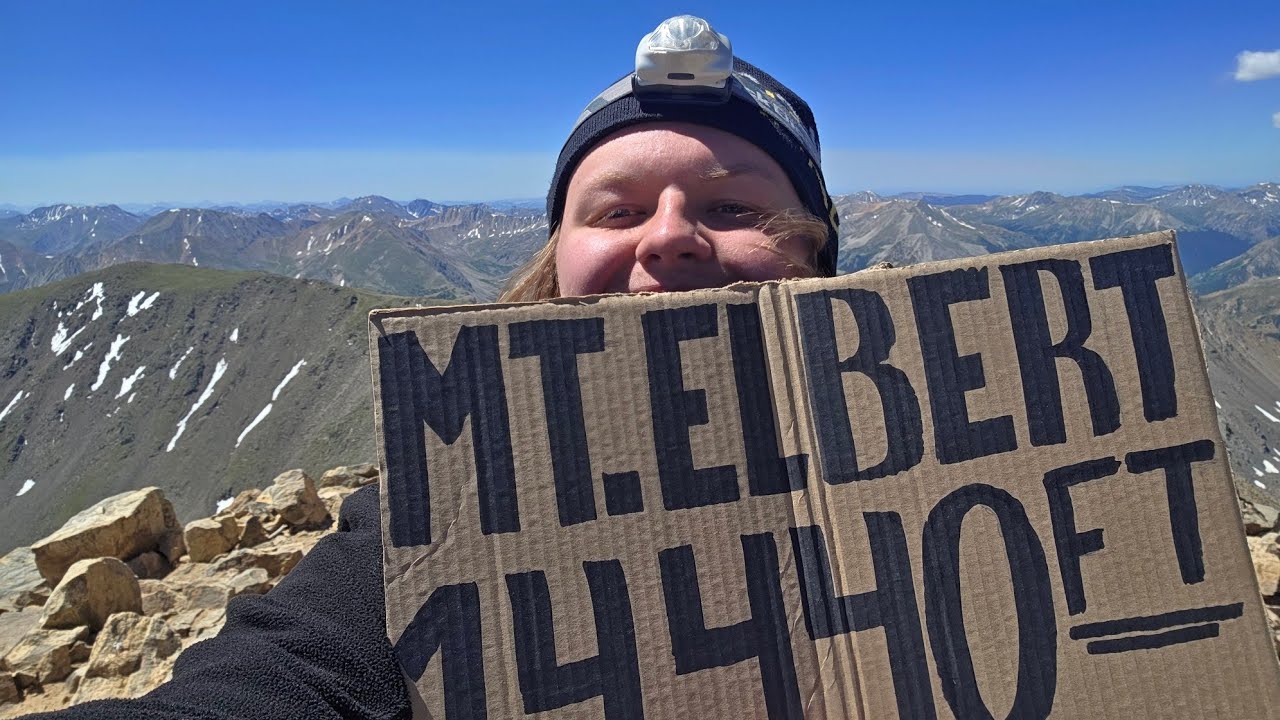 Finn grinning behind a cardboard sign reading "Mt. Elbert 14,440 ft" on the rocky summit