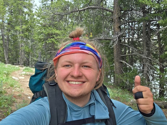 Finn in a tie-dye headband grinning with a thumbs up in the forest