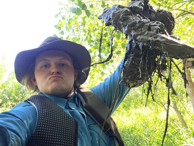 Finn in a sun hat holding up a muddy branch by the river